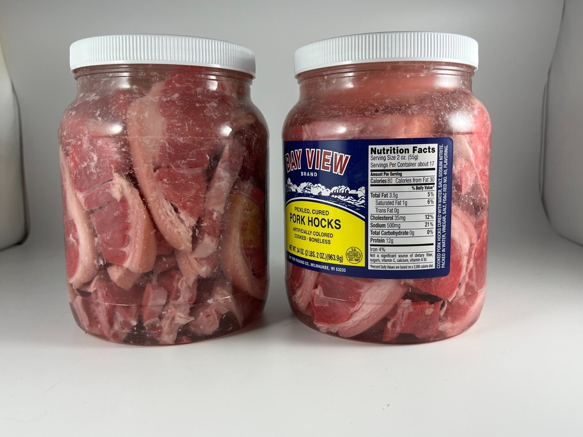A clear glass jar filled with pink pickled pork cubes in a pale vinegar brine with garlic cloves and black peppercorns, sitting on a bright kitchen counter