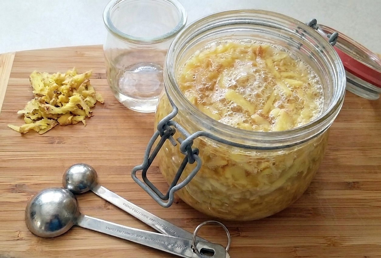 A clear glass jar of active ginger bug starter with visible bubbles and sliced ginger on a bright kitchen counter