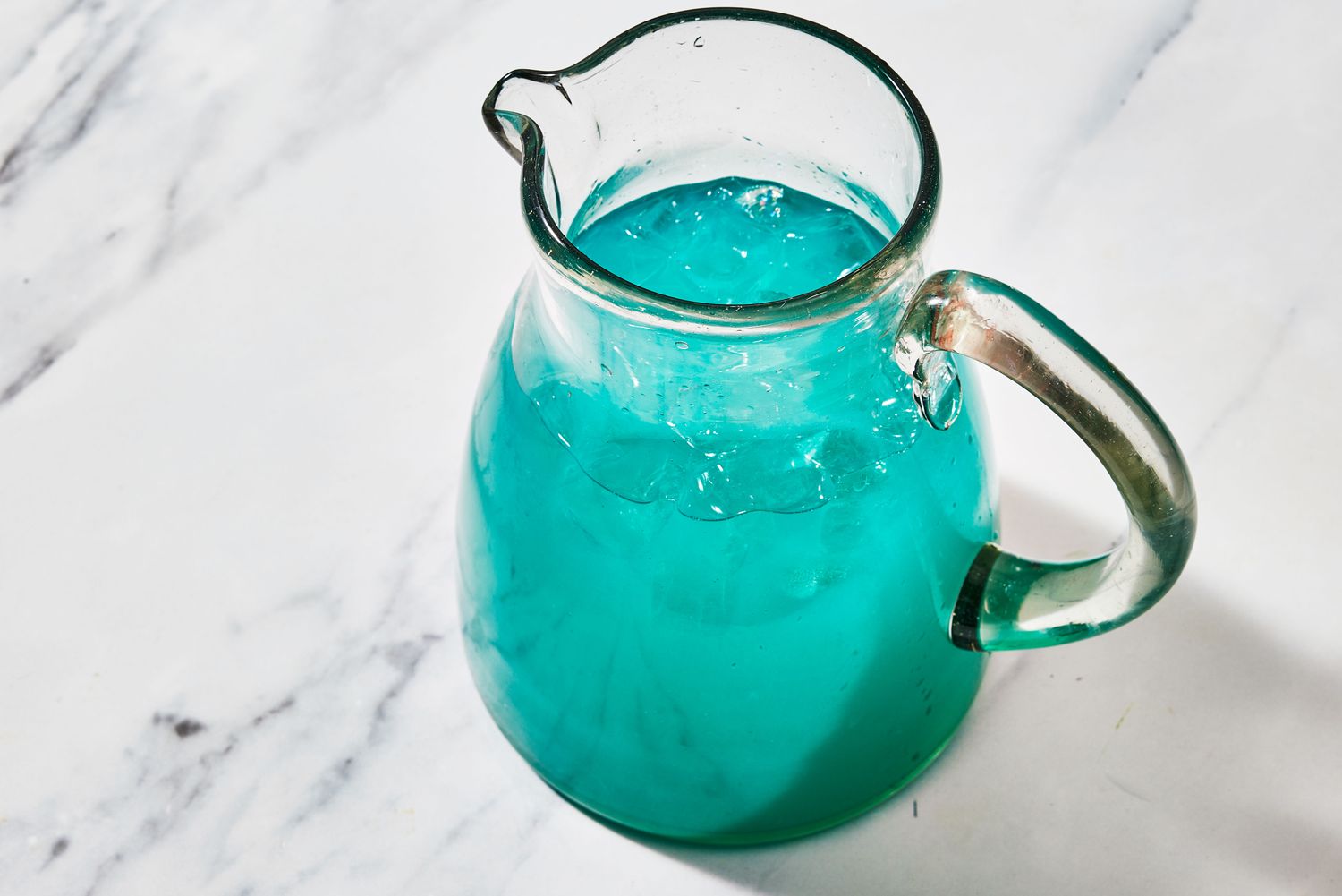 A clear glass pitcher filled with bright ocean-blue punch with ice, lime wheels, and pineapple wedges on a kitchen counter