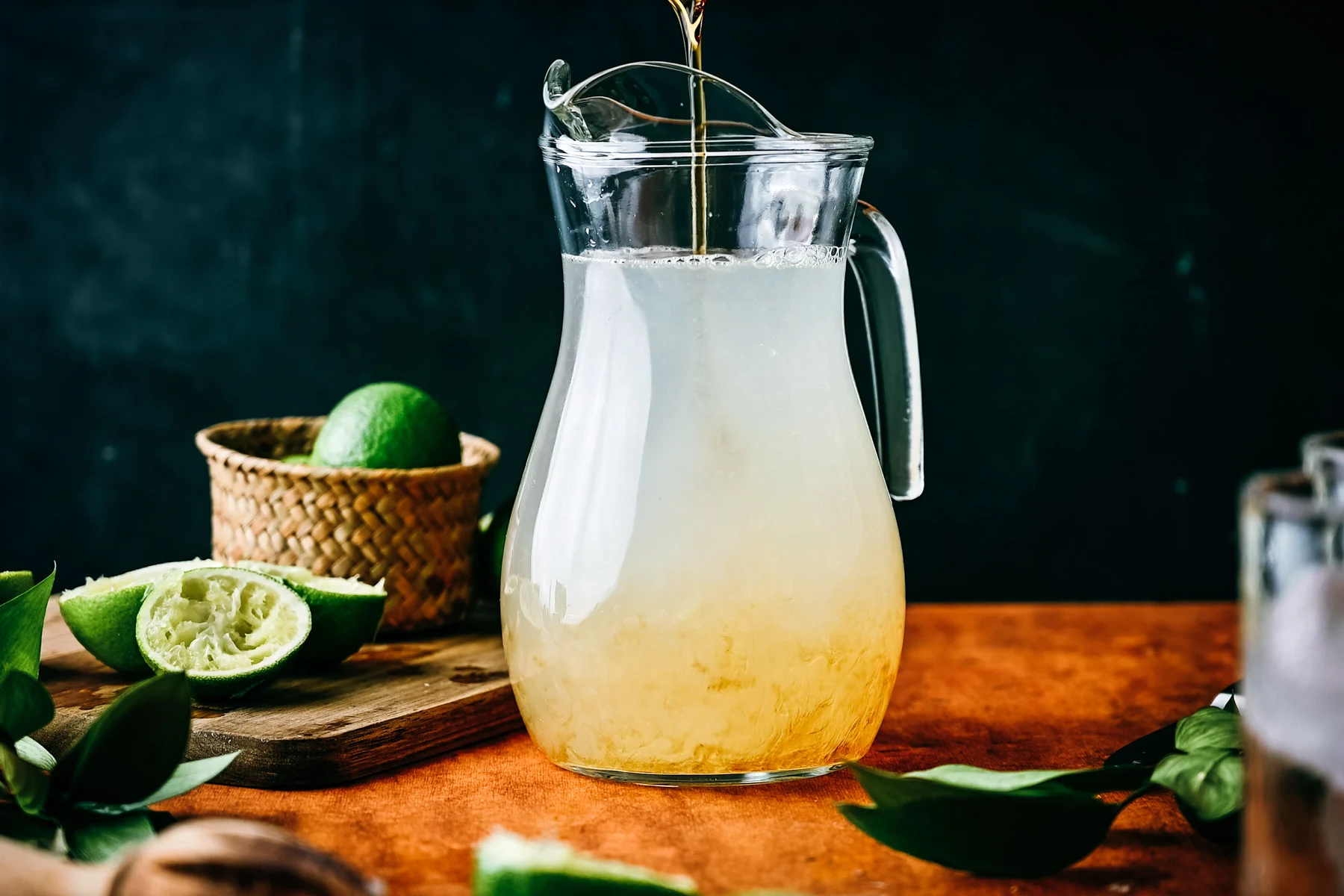 A clear glass pitcher of pale green homemade limeade with lime wheels and lots of ice on a bright kitchen counter, with two chilled glasses nearby
