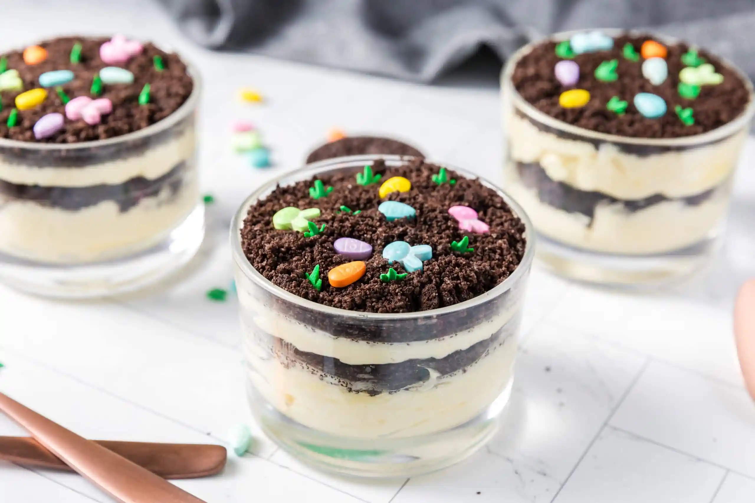 A clear glass trifle dish filled with layers of chocolate pudding and fluffy cream, topped with crushed chocolate sandwich cookie crumbs and a few gummy worms, set on a bright kitchen counter with soft natural light