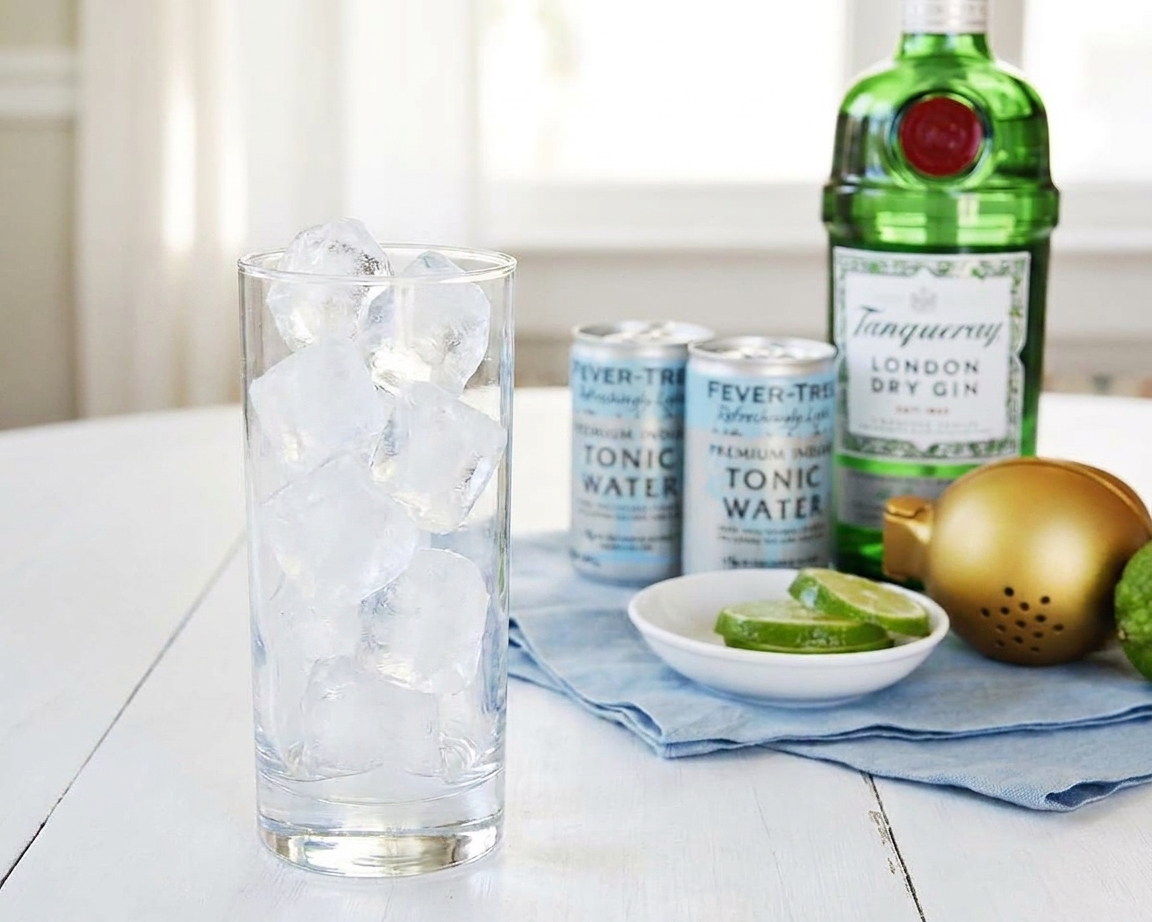 A clear highball glass being filled to the top with large ice cubes on a kitchen counter