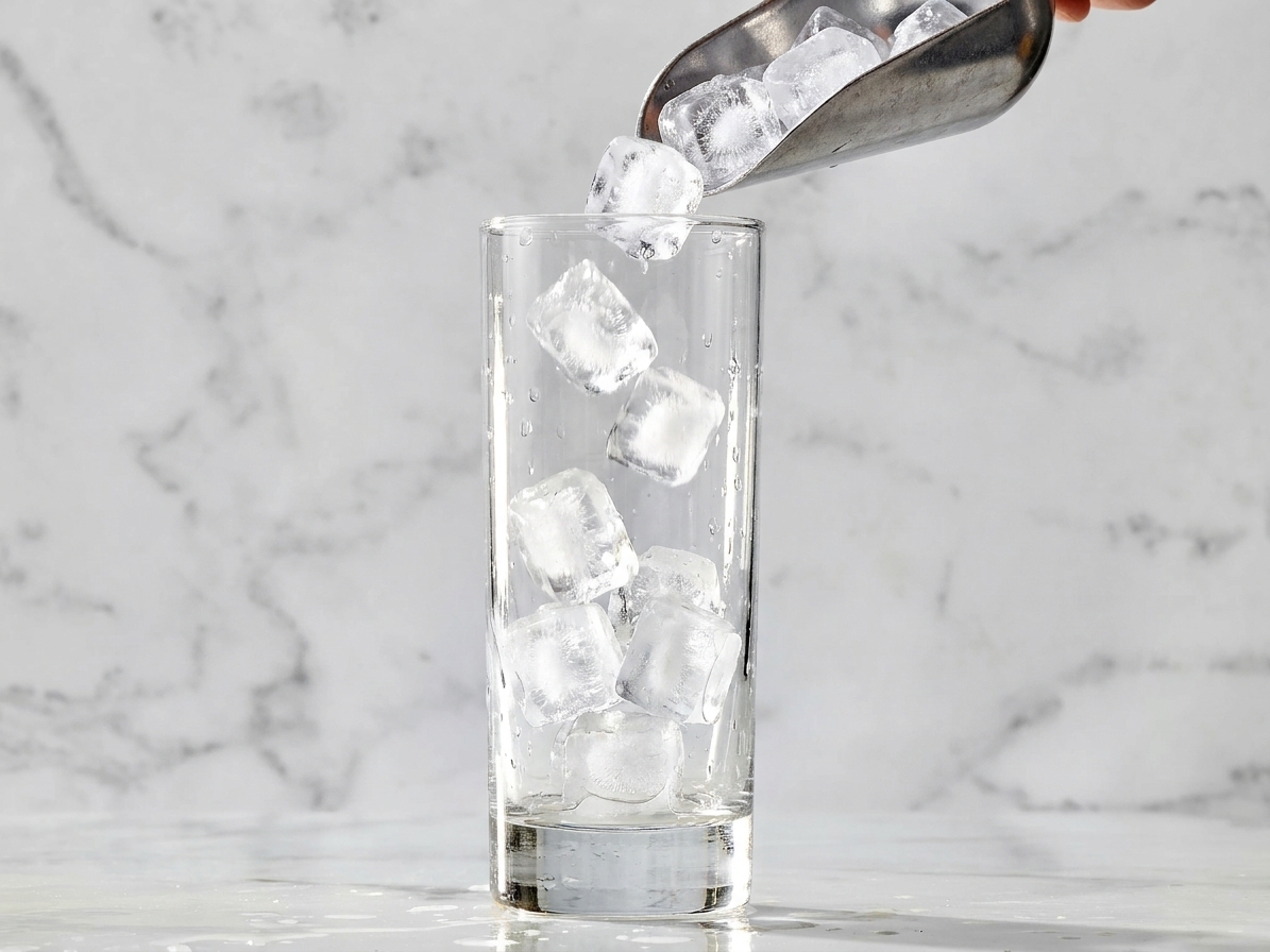 A clear highball glass being filled with fresh ice cubes on a clean kitchen counter