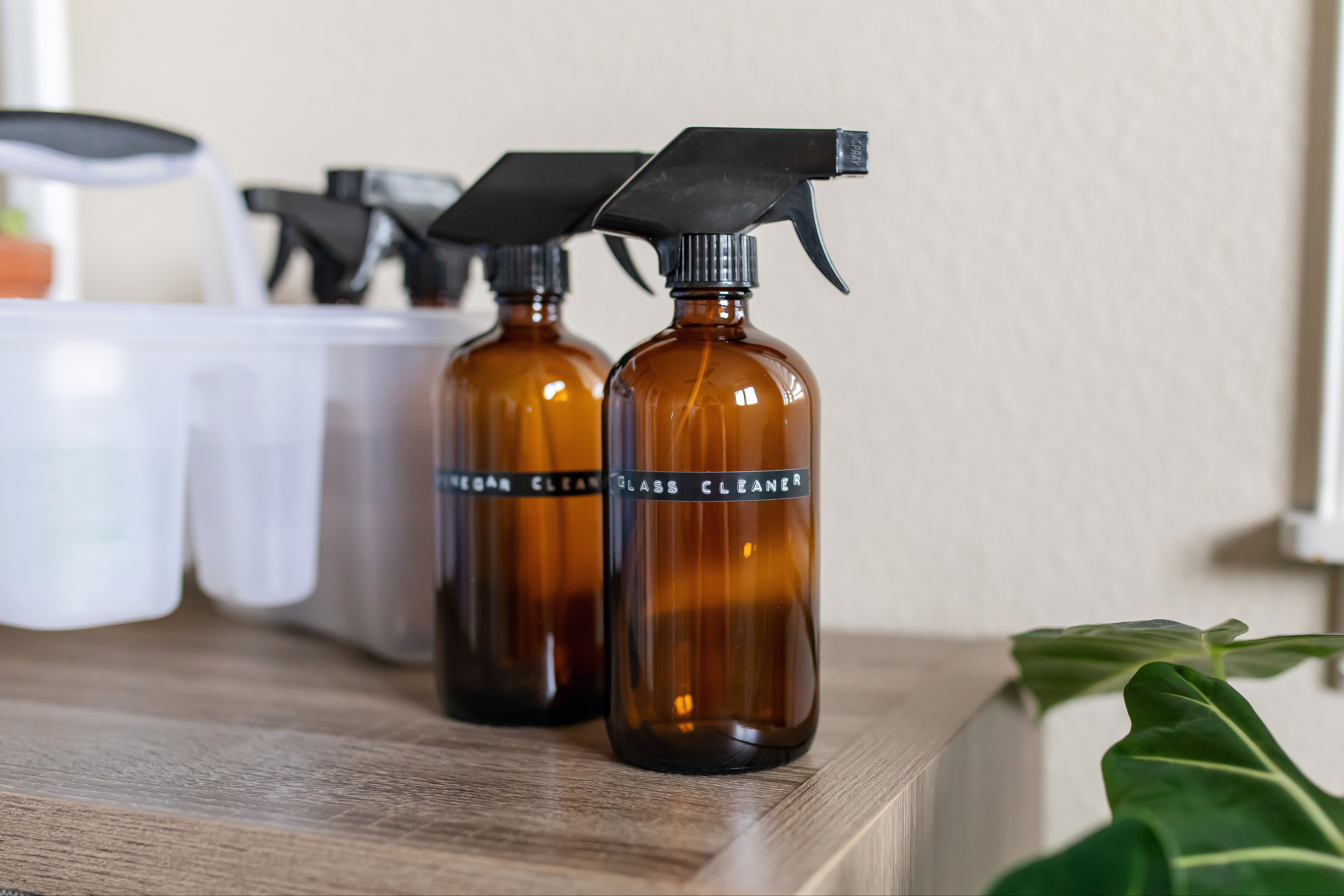 A clear spray bottle of homemade window cleaner on a bright kitchen counter beside a microfiber cloth and a roll of paper towels, natural morning light