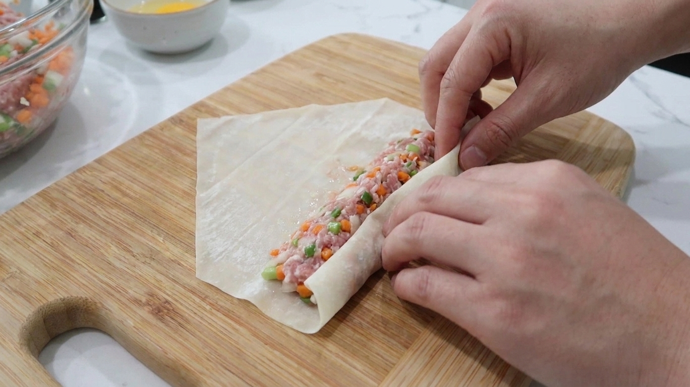 A close-up of a lumpia wrapper being rolled tightly around a thin line of pork filling on a wooden cutting board