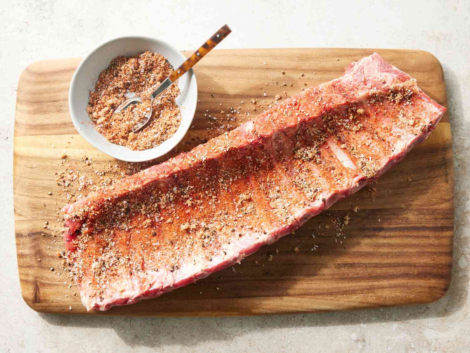 A close-up of a small bowl of smoky spice rub next to a raw pork butt roast on a cutting board