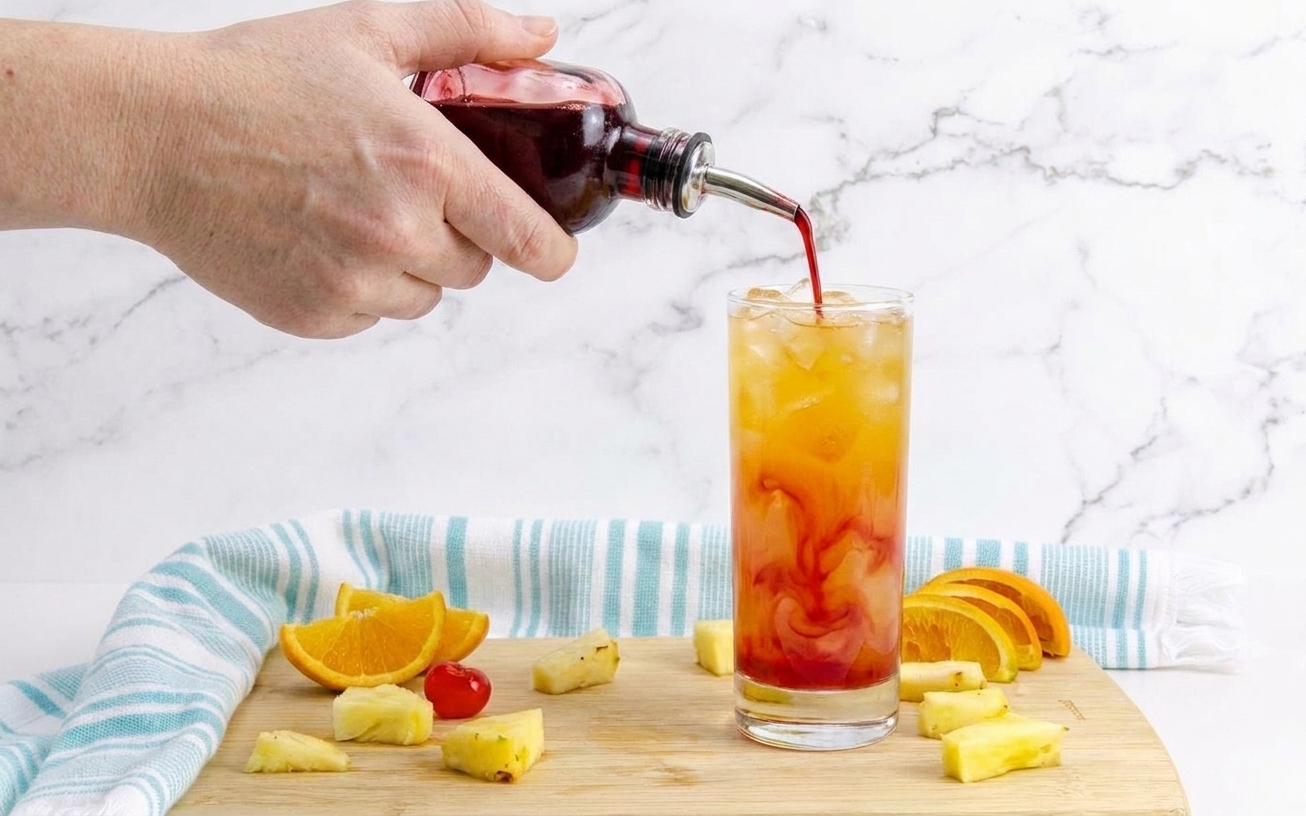 A close-up of grenadine being poured into a glass of tropical rum cocktail, creating a red swirl through the ice