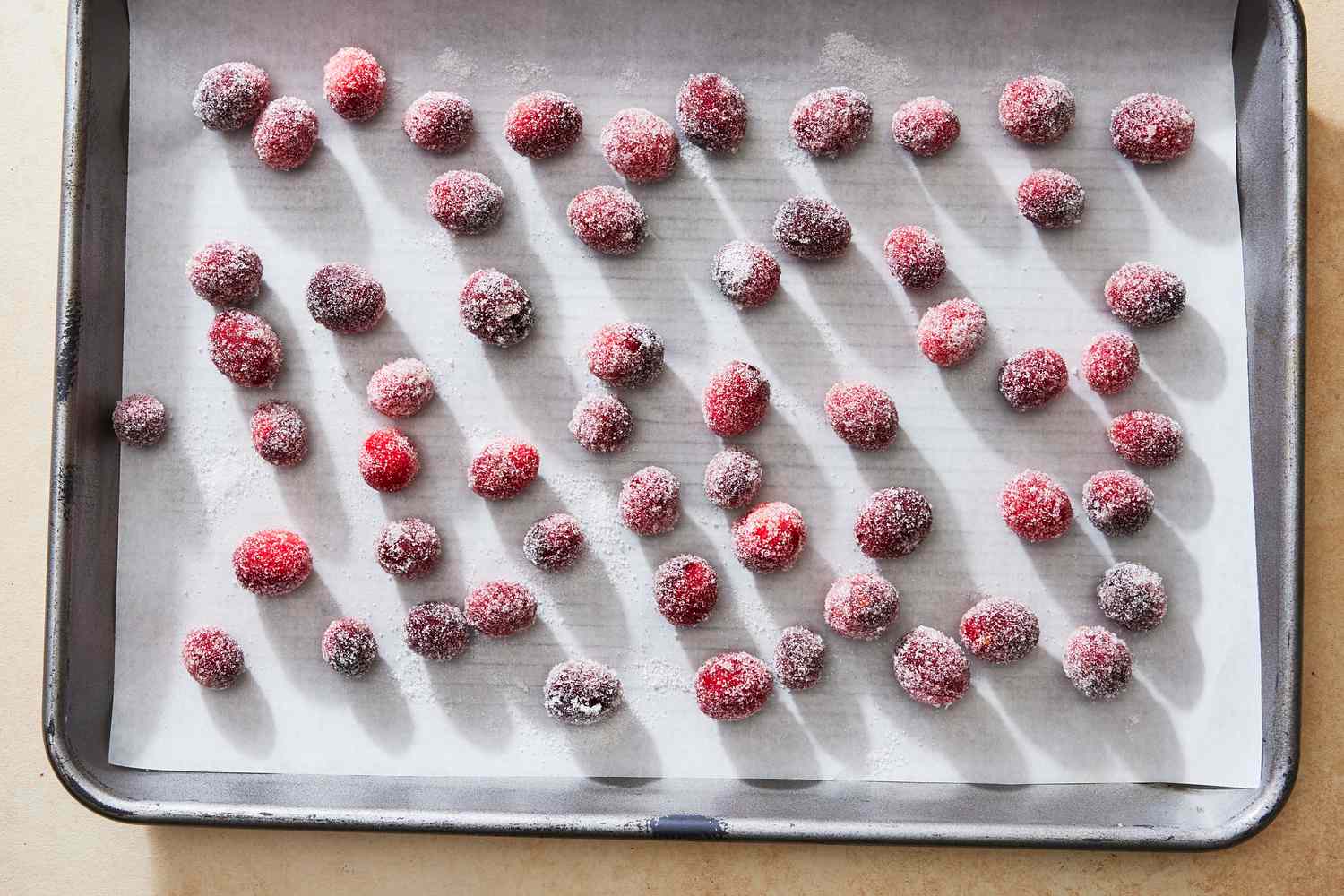 A close-up of sparkling sugared cranberries piled on parchment paper on a warm kitchen counter, with a small bowl of sugar nearby