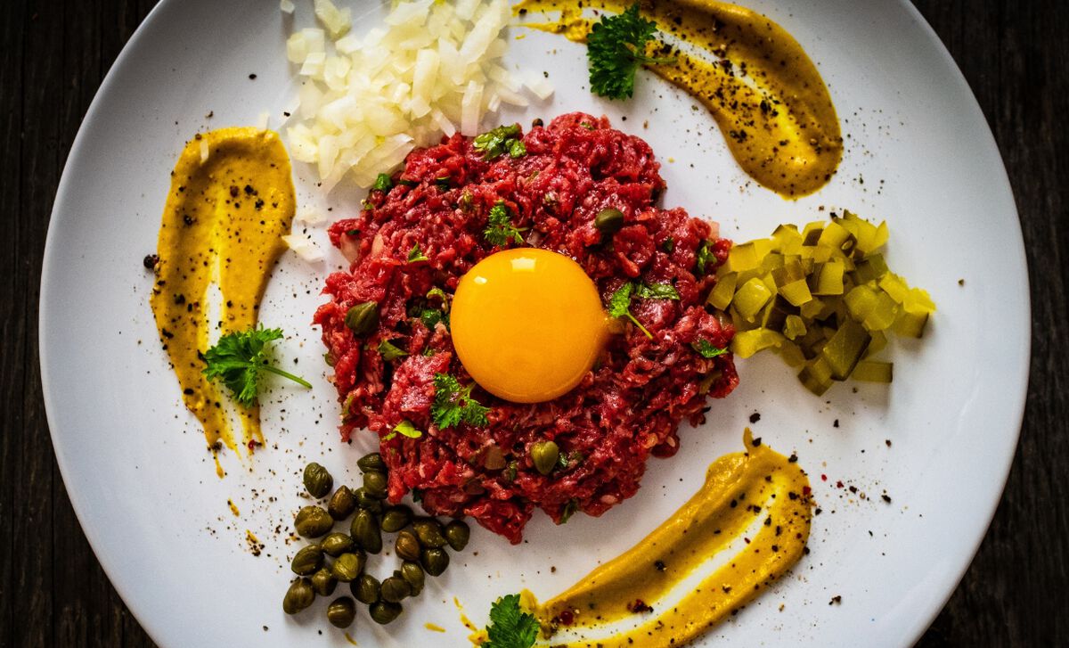 A close-up of steak tartare on a plate with a raw egg yolk nestled on top and toasted bread slices nearby on a kitchen counter