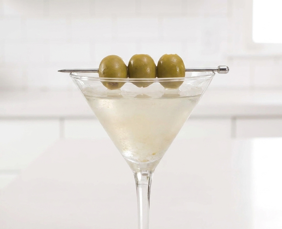 A close-up of three green olives on a cocktail pick resting across the rim of a cloudy extra dirty martini