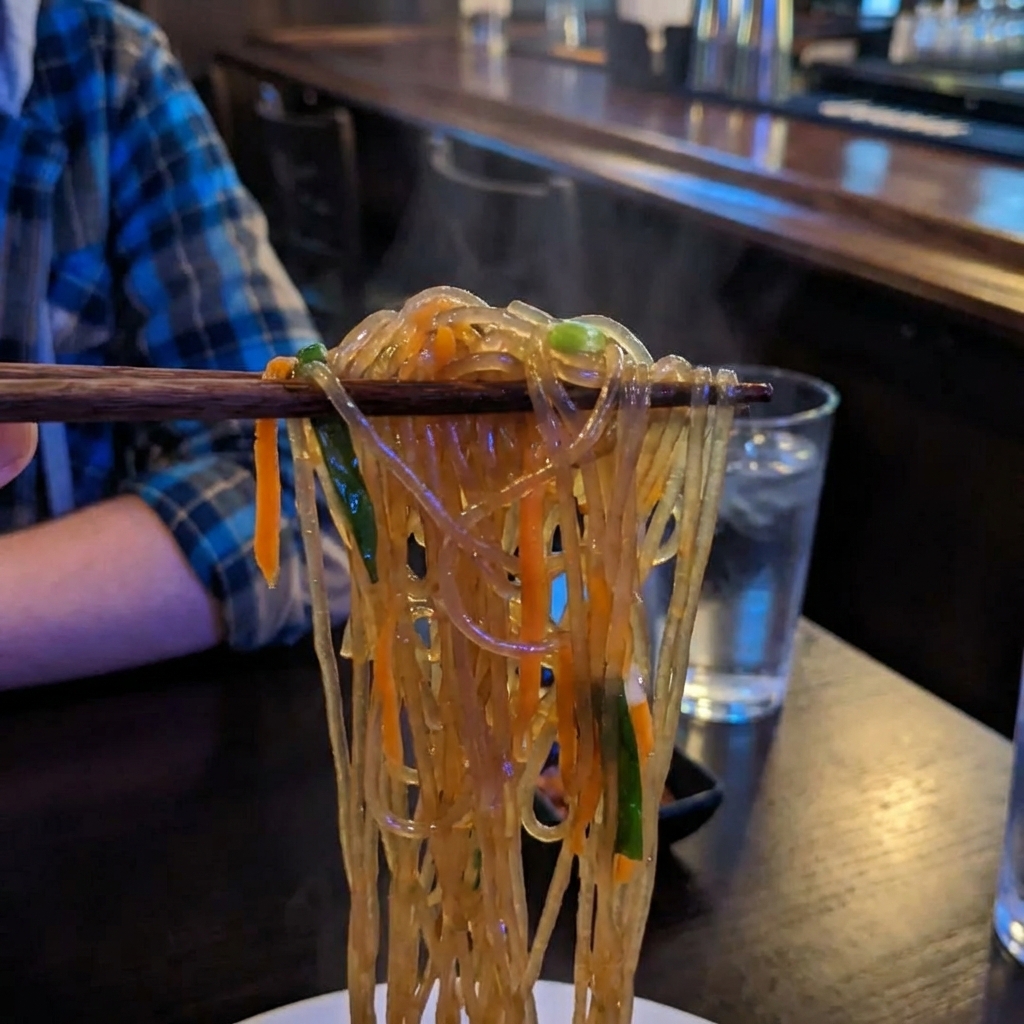 A close up photo of chopsticks lifting a tangle of glossy japchae noodles with strips of carrot and scallions