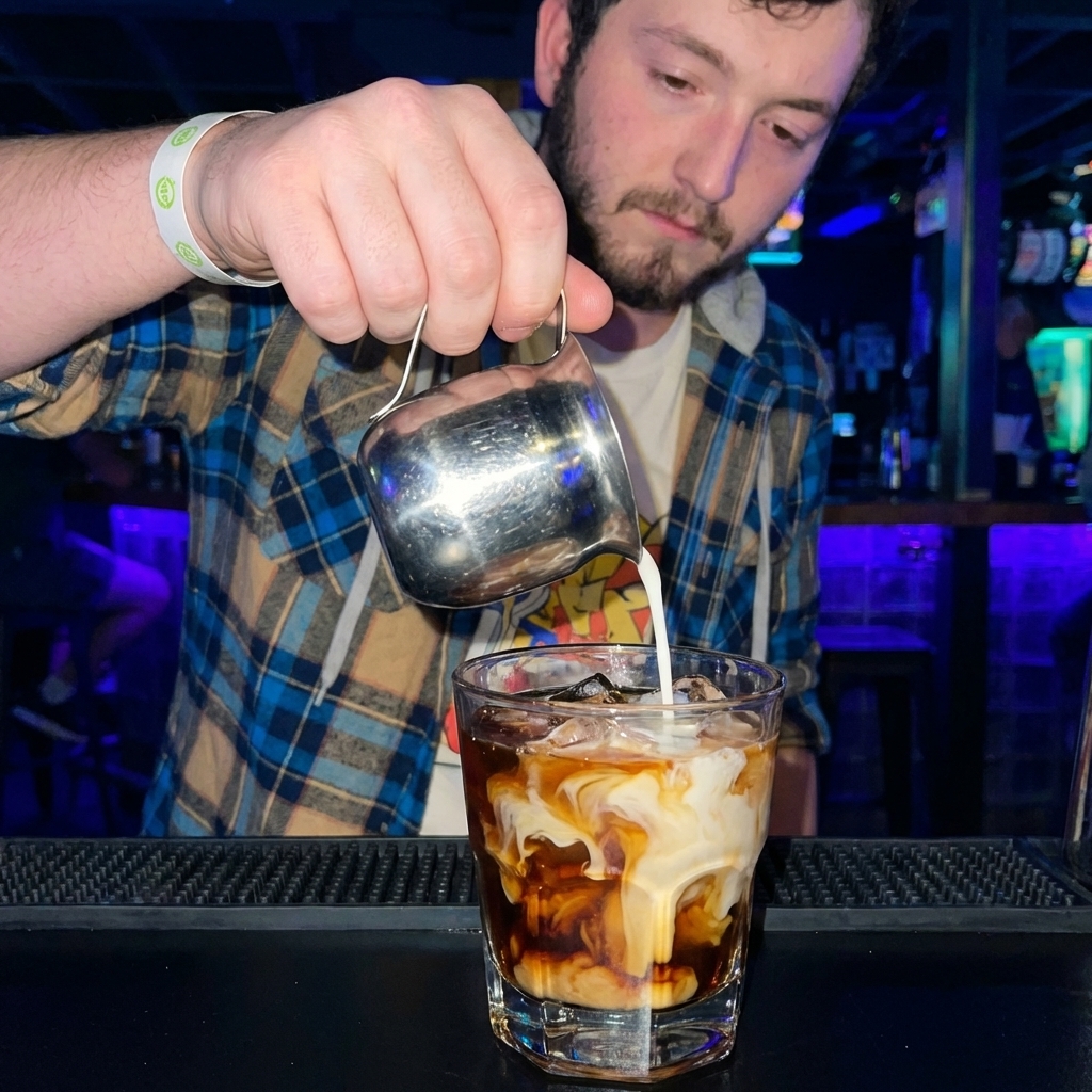 A close-up photo of cream being poured into a rocks glass of vodka and coffee liqueur over ice, creating a marbled swirl