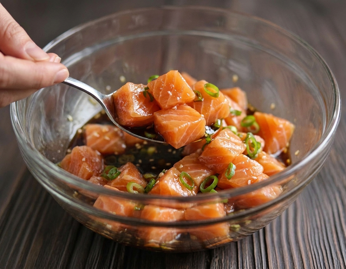 A close-up photo of cubed salmon in a glass bowl being tossed with soy sauce and sesame oil marinade with sliced scallions