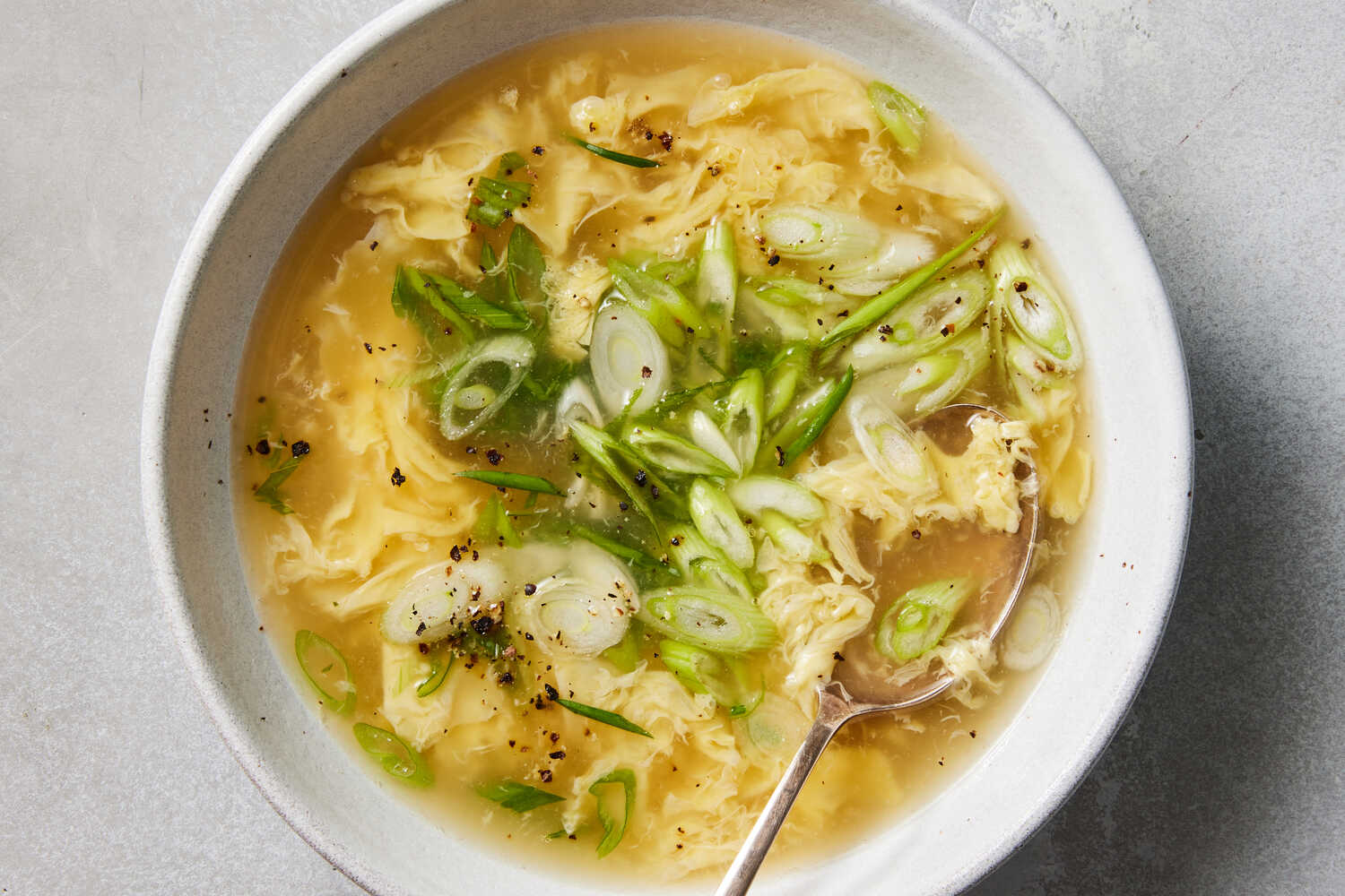 A close-up photo of egg drop soup with delicate egg ribbons and green onion slices floating on the surface in a white bowl