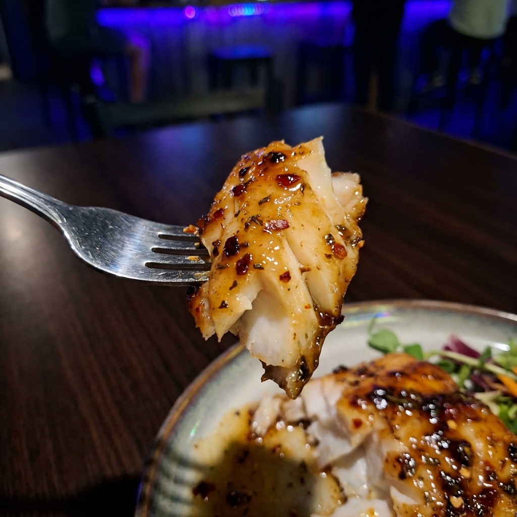 A close-up photograph of a fork lifting a flaky piece of baked cod with glossy glaze and chili flakes