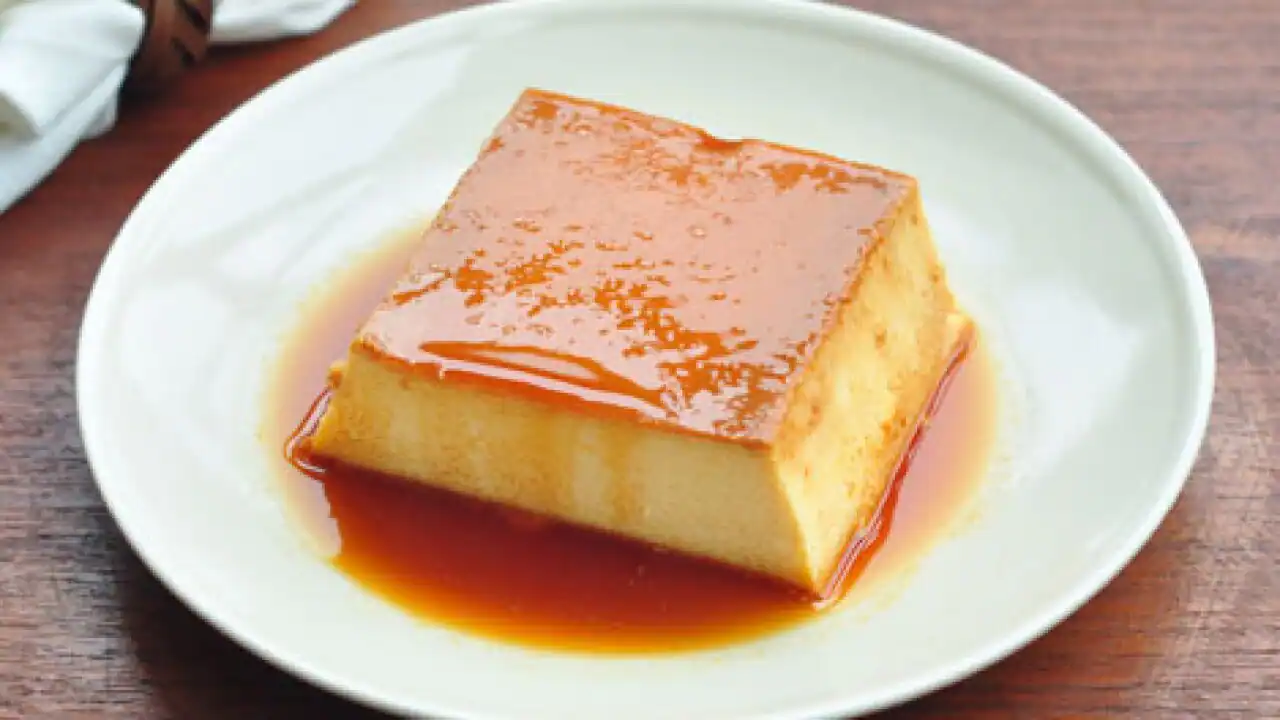 A close-up slice of leche flan showing a smooth, creamy custard interior and a thin dark caramel layer on top with caramel sauce on the plate