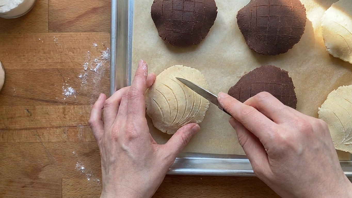 A close view of a concha topping being scored with a small knife into curved shell-like lines on a baking sheet