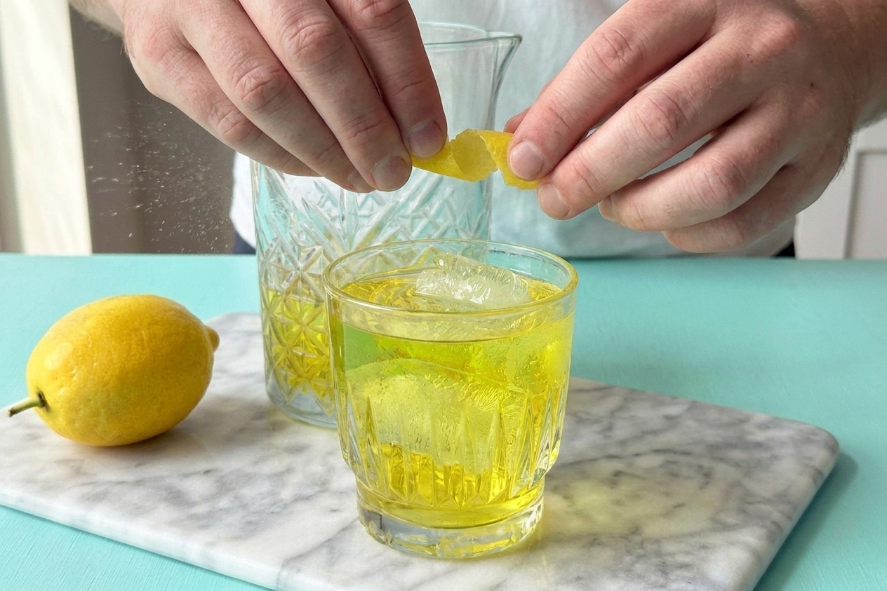 A close view of a lemon peel being twisted over a rocks glass to release citrus oils onto a chilled White Negroni