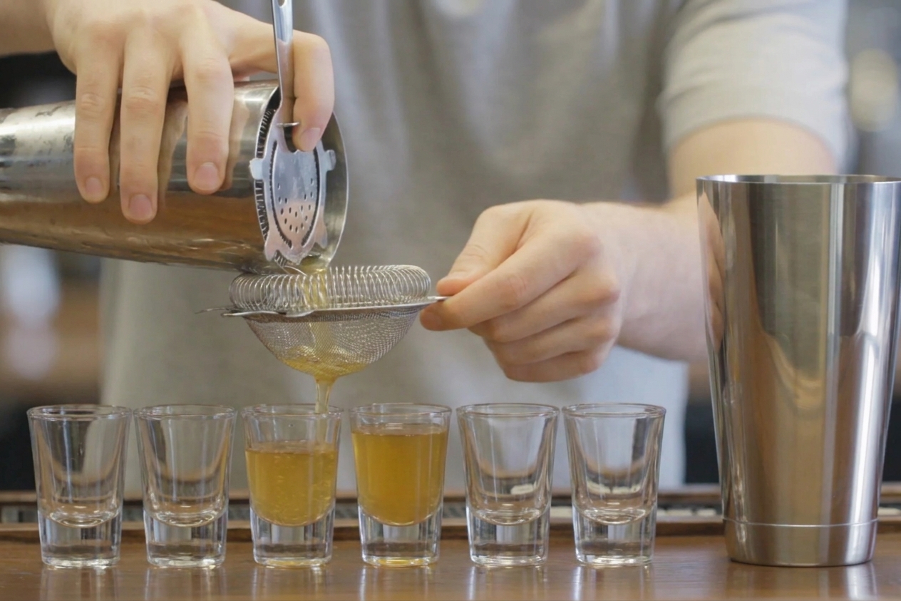 A cocktail being strained from a shaker into a row of shot glasses on a kitchen counter