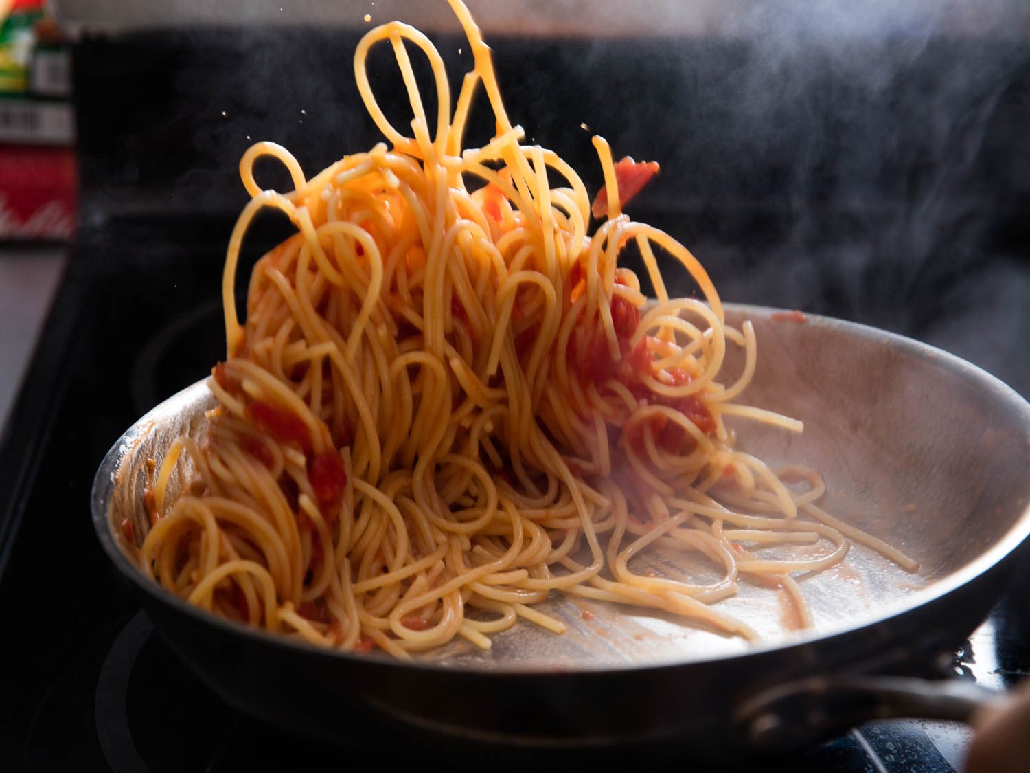 A cook using tongs to toss saucy Chinese noodles in a large skillet on a home stove with steam rising