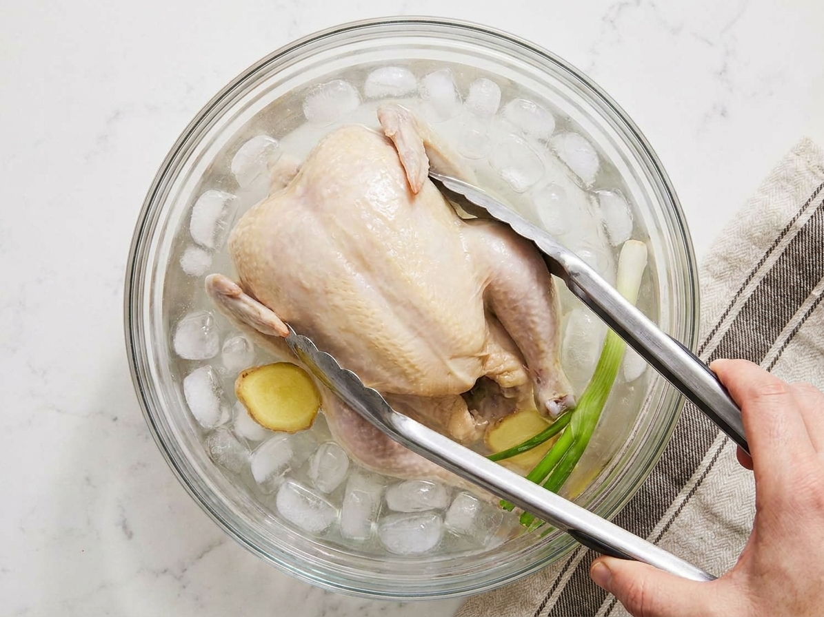 A cooked poached chicken being lowered into a bowl of ice water in a home kitchen, with tongs holding it