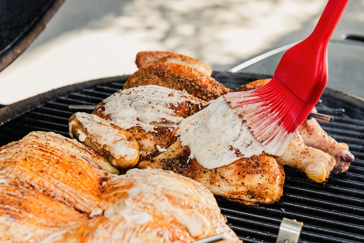 A cook's hands basting chicken thighs on a hot grill with a silicone brush, with visible caramelized edges and a small pot of butter baste nearby