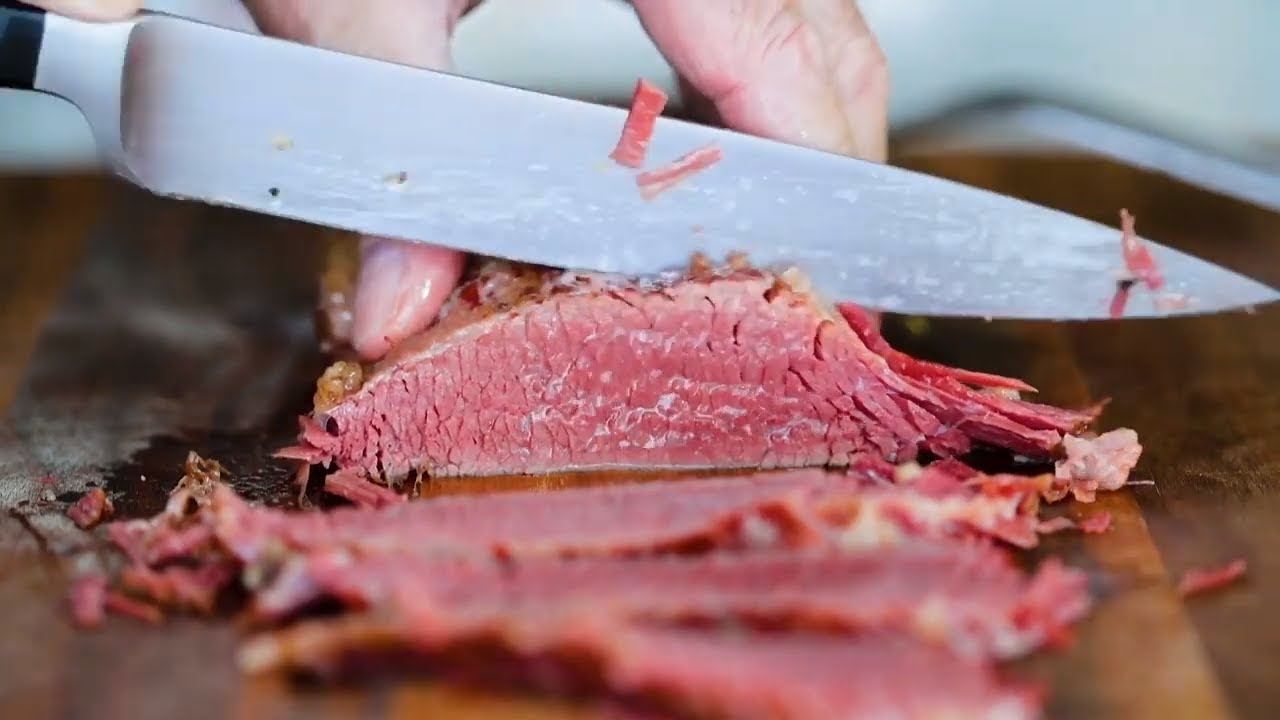 A cook’s hands slicing rested corned beef brisket against the grain on a wooden cutting board, with cabbage and potatoes waiting nearby