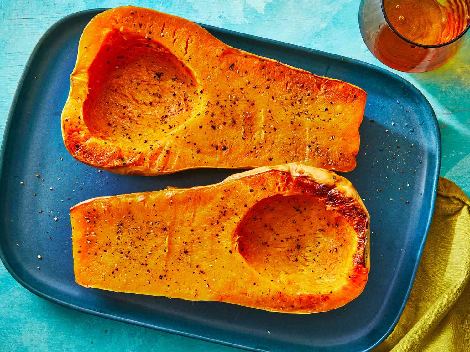 A cook’s hands sprinkling cracked black peppercorn and salt over butternut squash cubes on a sheet pan on a home kitchen counter