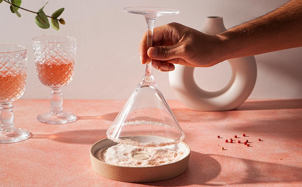 A coupe glass being rimmed with a lemon wedge next to a small plate of sugar on a kitchen counter