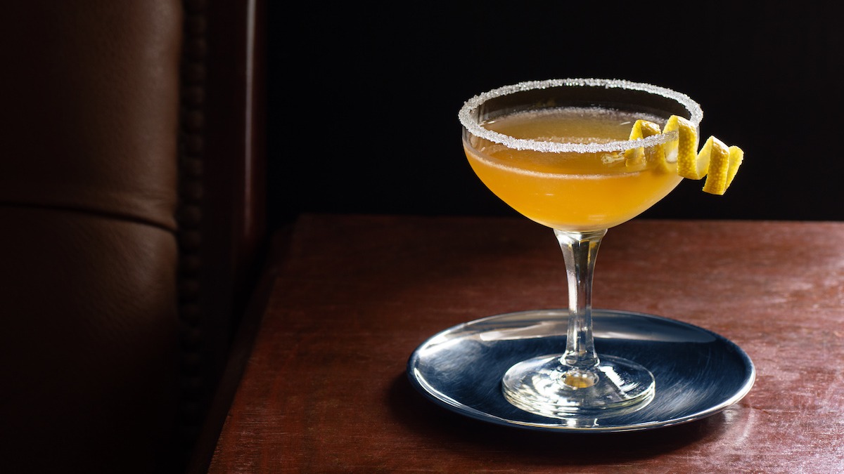 A coupe glass being rimmed with lemon juice and dipped into sugar on a small plate on a kitchen counter