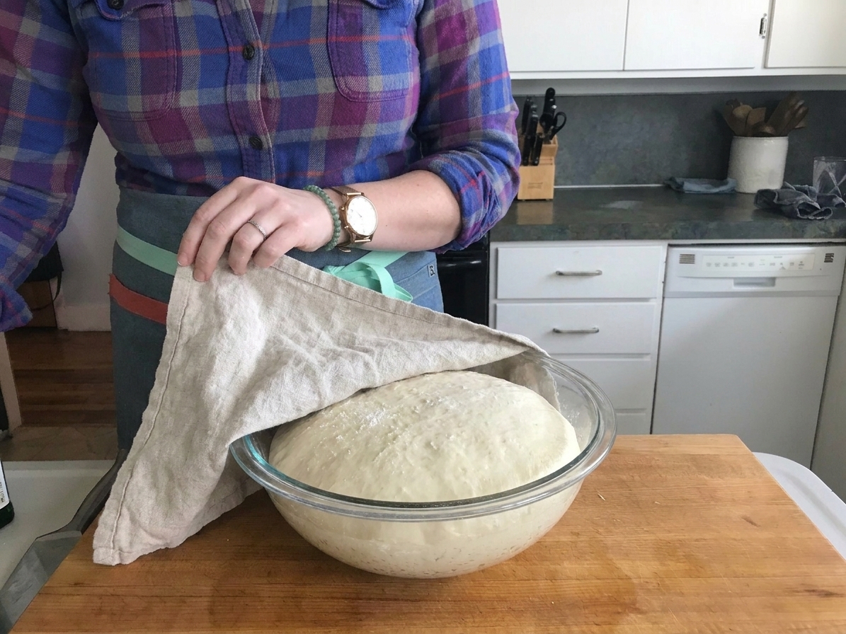 A covered bowl of pizza dough mid-rise on a countertop, dough visibly puffed and airy under a towel