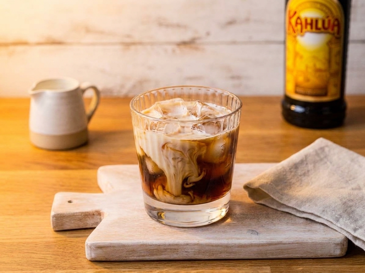 A creamy White Russian cocktail in a rocks glass with large clear ice cubes on a warm-toned kitchen counter, soft evening lighting