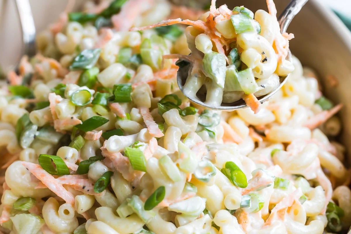 A creamy scoop of Hawaiian macaroni salad sprinkled with black pepper on a plate beside simple barbecue-style chicken and rice, bright natural kitchen light
