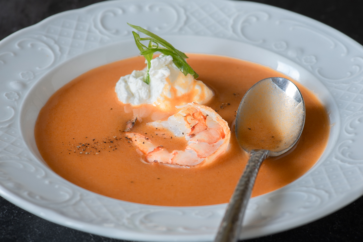 A creamy shrimp bisque in a white bowl topped with seared shrimp and chopped chives on a wooden table with a spoon beside it