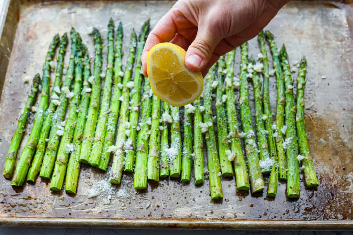 A cutting board on a kitchen counter with fresh asparagus, a lemon, garlic cloves, Parmesan, olive oil, salt, pepper, and red pepper flakes arranged for cooking