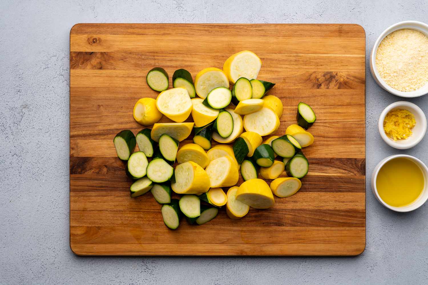 A cutting board on a kitchen counter with neatly sliced zucchini and yellow squash coins, a chef knife, and a small bowl for scraps