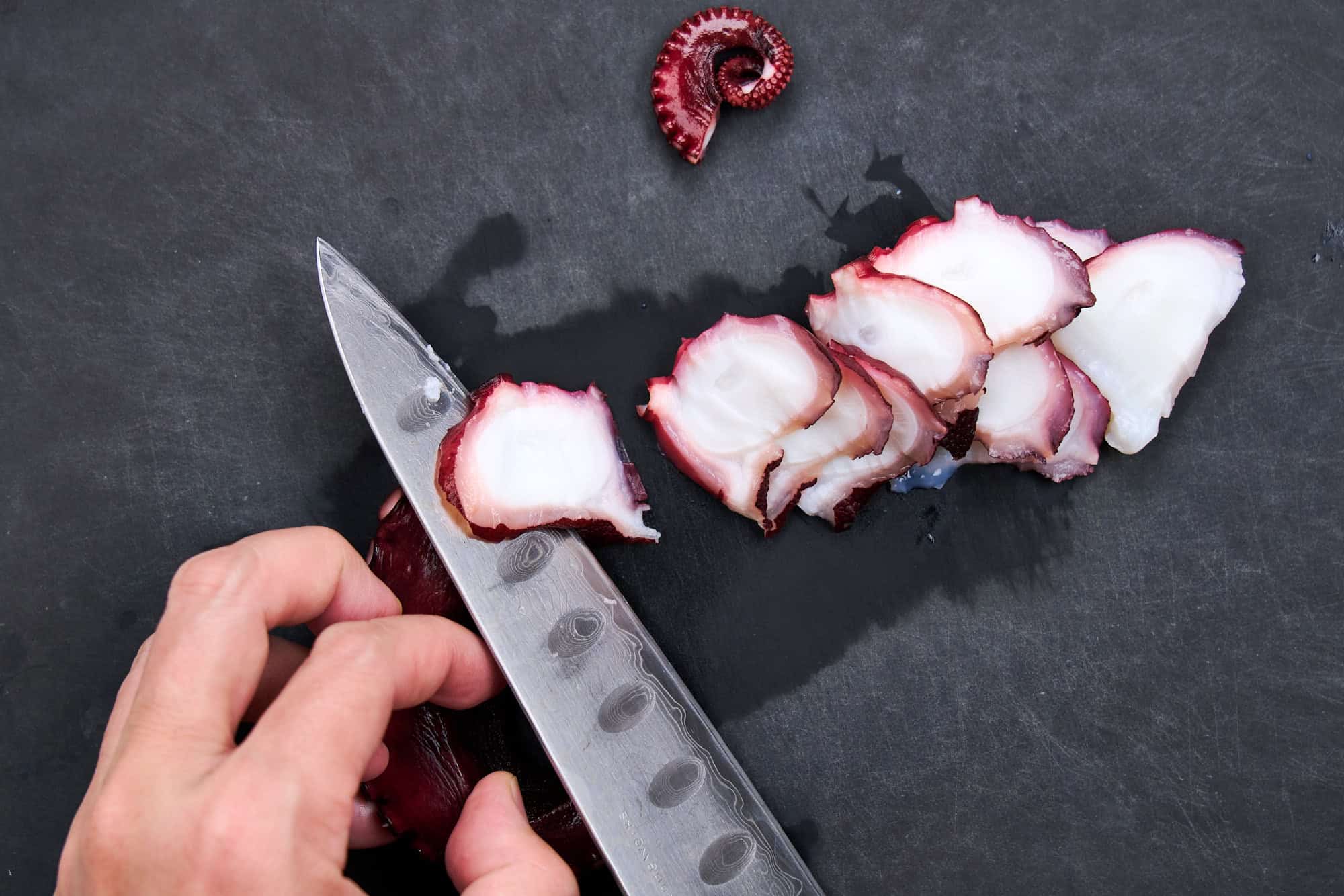 A cutting board with a cooked octopus tentacle being sliced into coins with a chef's knife under warm kitchen lighting