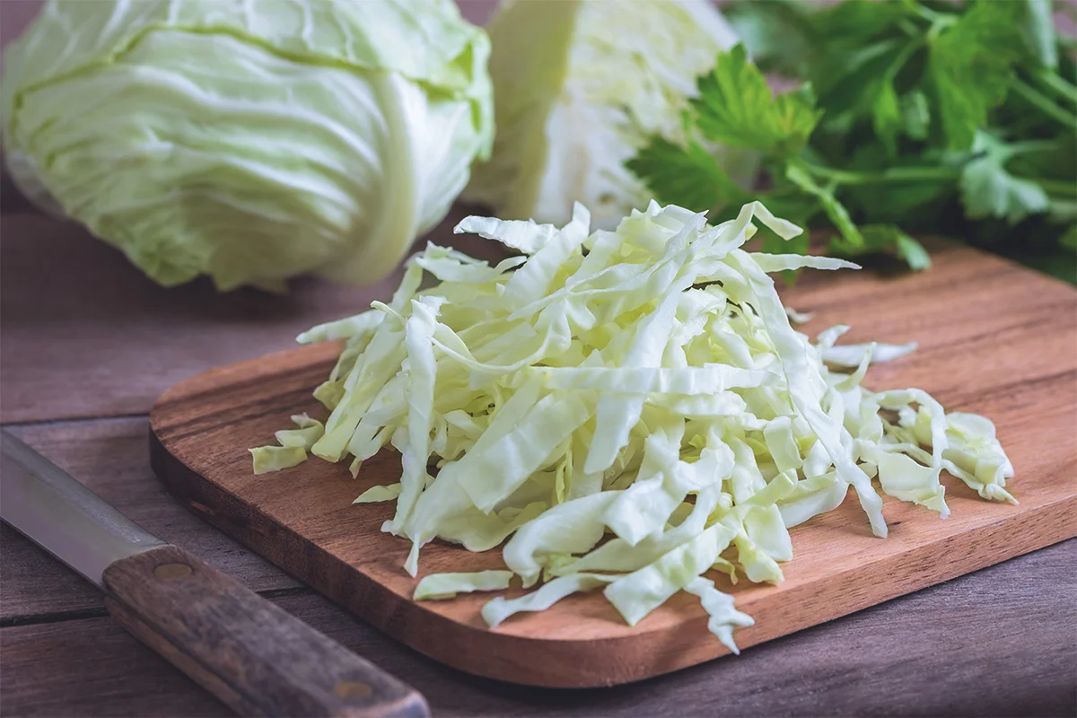 A cutting board with a pile of chopped green cabbage and a chef's knife on a bright kitchen counter