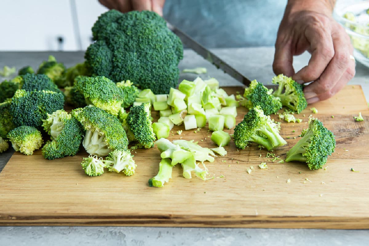 A cutting board with broccoli being chopped into small florets with a chef’s knife on a bright kitchen counter