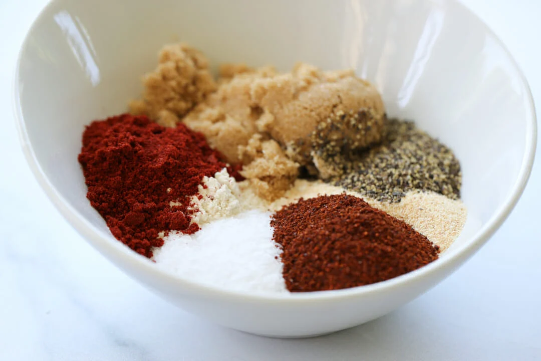 A cutting board with chopped onion and minced garlic beside small bowls of smoked paprika, oregano, and red pepper flakes on a kitchen counter