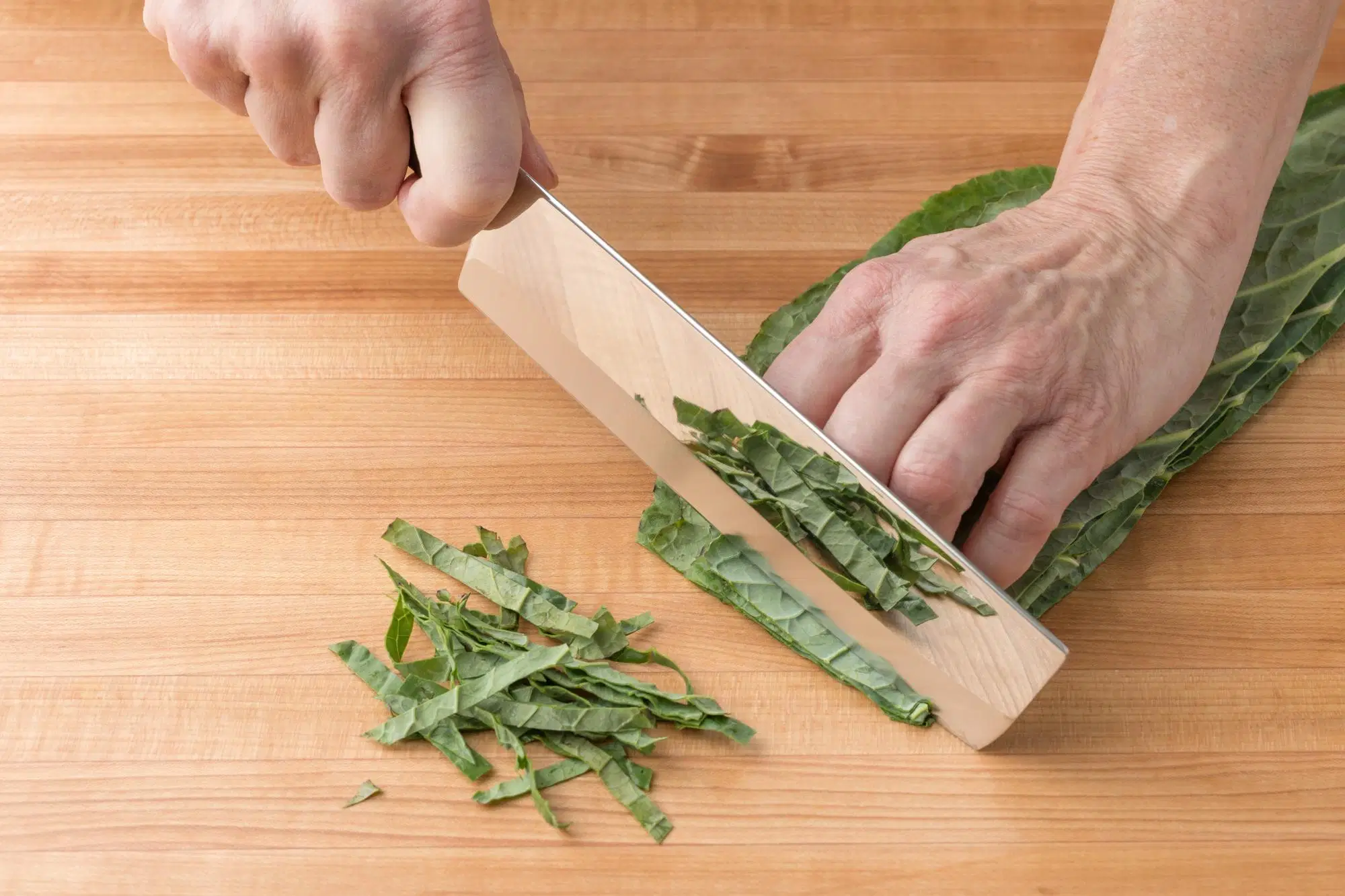 A cutting board with collard greens being chopped into ribbons with a chef’s knife, with a bowl nearby