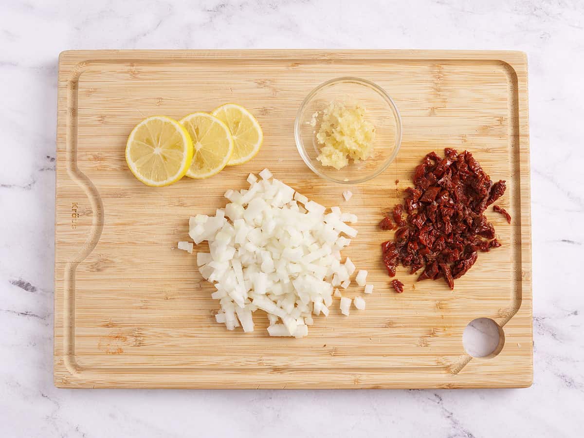A cutting board with diced onion, minced garlic, and a zested lemon beside a chef's knife