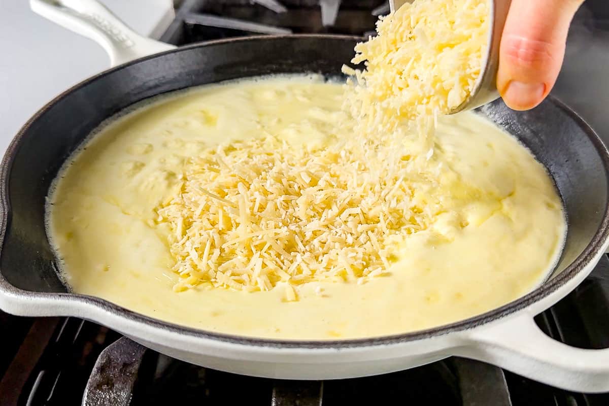 A cutting board with finely grated Parmesan, minced garlic, and cubes of butter prepped beside a skillet on a home kitchen counter