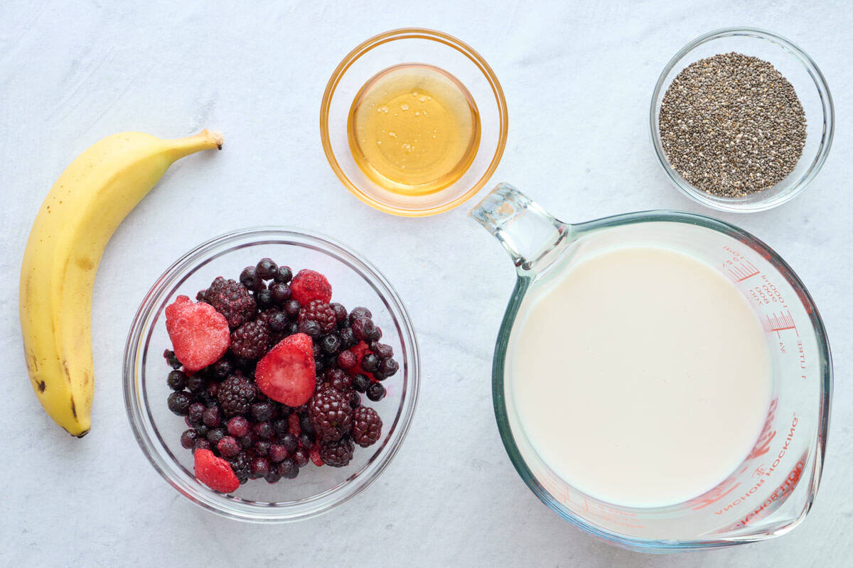 A cutting board with frozen mixed berries in a bowl, half a banana, and a small dish of chia seeds with a spoon