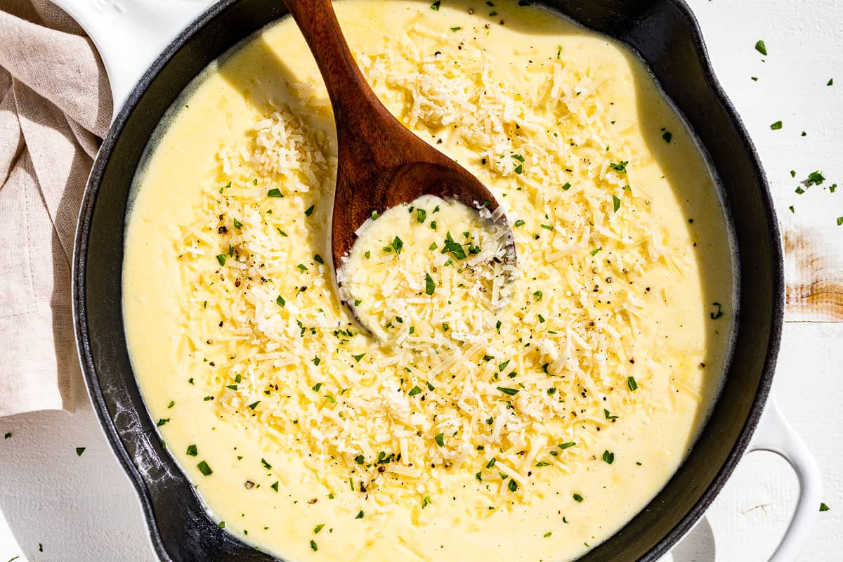 A cutting board with minced garlic, a bowl of finely grated Parmesan, a small dish of butter, and a measuring cup of heavy cream on a kitchen counter