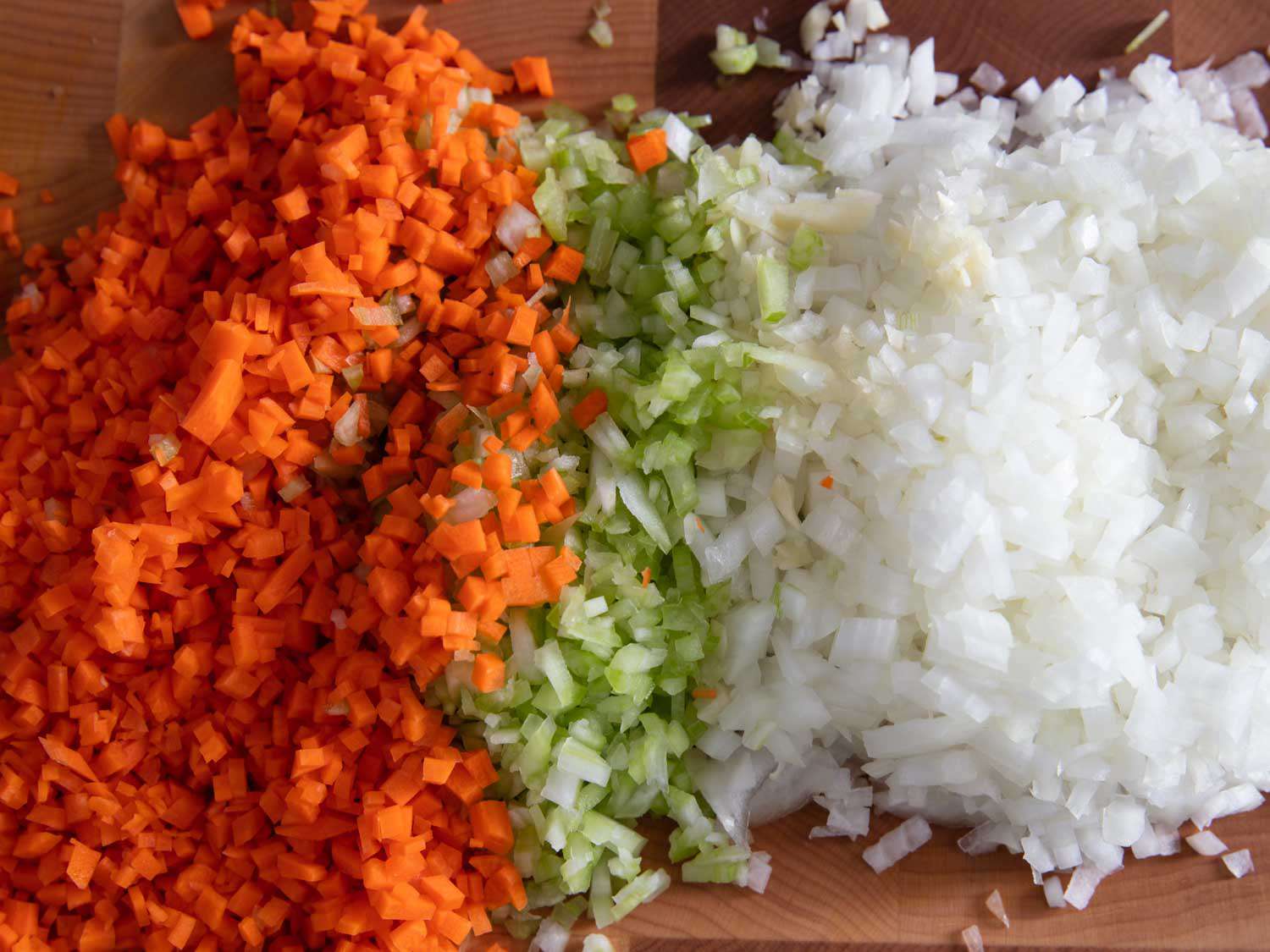 A cutting board with neatly chopped carrots, celery, and diced onion beside minced garlic in a bright home kitchen