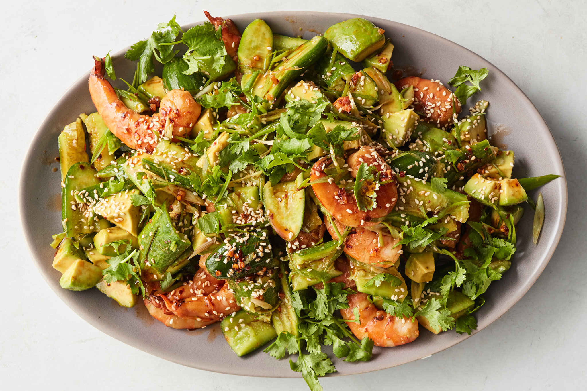 A cutting board with peeled cooked shrimp, diced avocado, chopped cucumber, sliced red onion, and fresh cilantro ready for Mexican shrimp cocktail prep on a kitchen counter