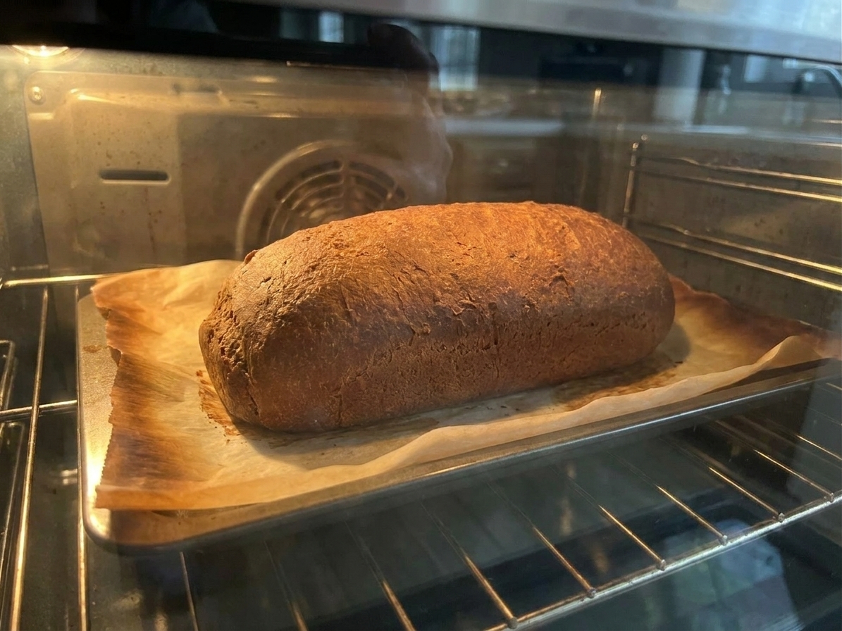 A dark rye loaf baking on a parchment-lined sheet pan inside a home oven