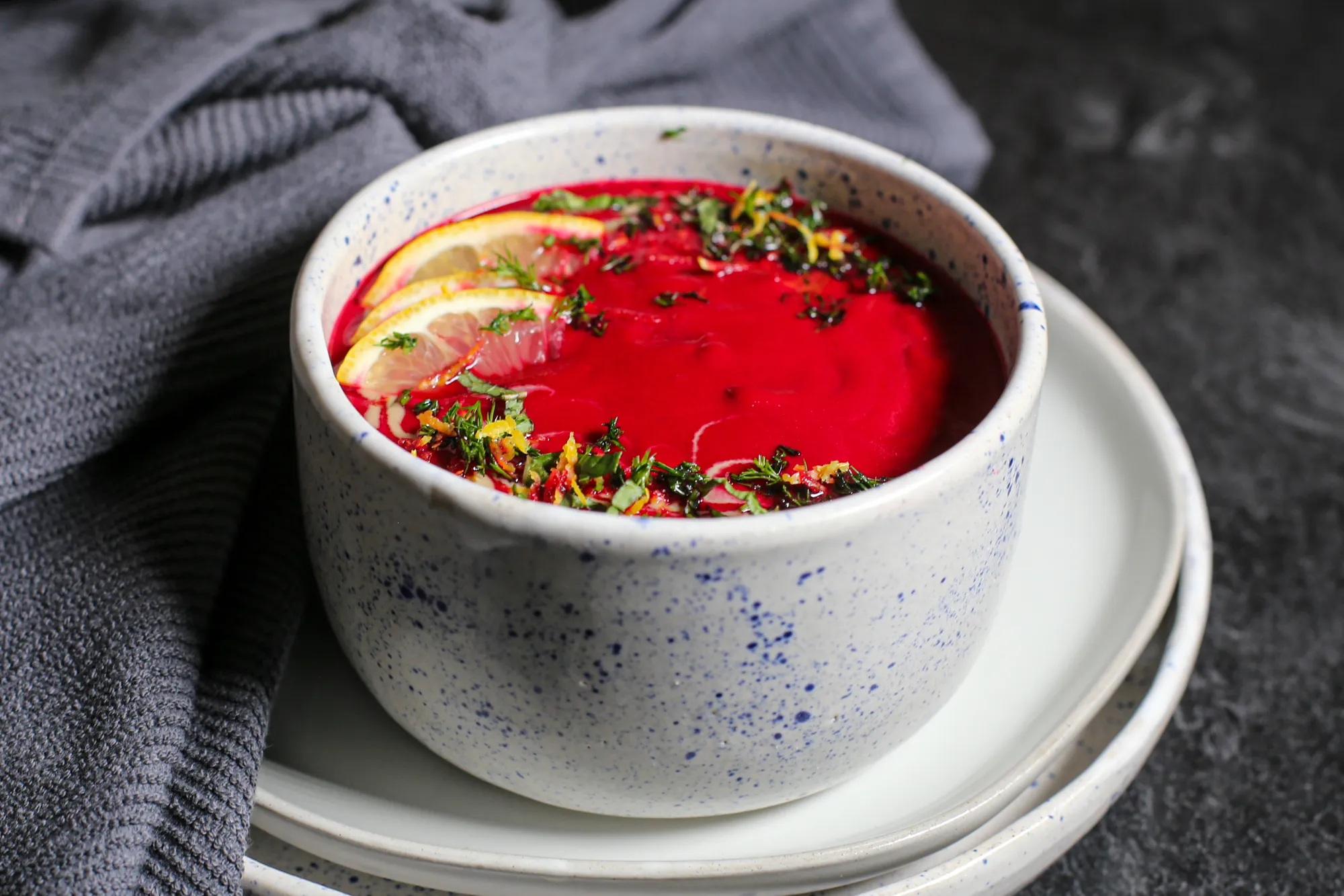 A deep magenta bowl of creamy beet soup topped with a swirl of yogurt, fresh dill, and cracked black pepper on a light stone kitchen table with a spoon and napkin