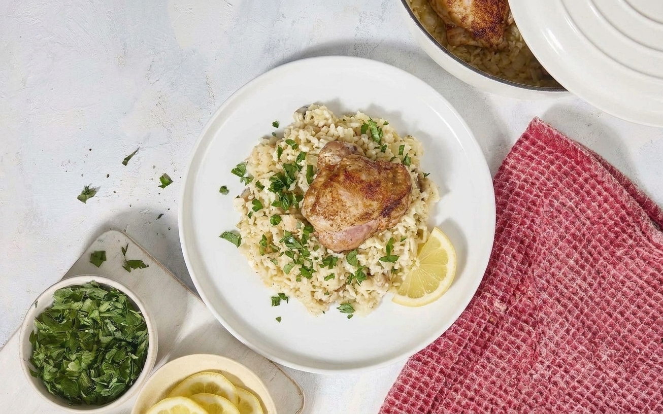 A dinner plate with a scoop of lemony rice topped with golden chicken and parsley, with a lemon wedge on the side on a simple table setting